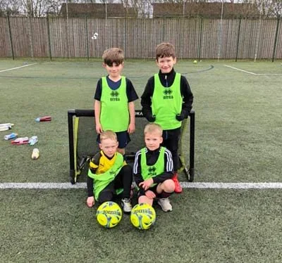 Young players in green bibs gathered round a small goal with footballs